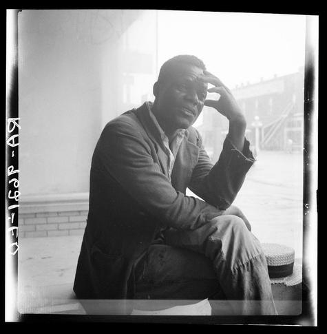 The photograph is a black and white image depicting an individual seated on the ground in front of what appears to be a building with brick walls. The person, whose gender isn't clearly identifiable from this angle but seems male based on clothing style, has one hand raised to his head as if scratching or shielding his eyes from sunlight, suggesting it might be outdoors and potentially sunny.

He is wearing a dark jacket over lighter-colored pants that are somewhat faded at the knees, indicating wear. His attire suggests casual or work-related activity rather than leisure. The person's expression seems contemplative or tired, with visible lines on his forehead adding to this impression of fatigue or deep thought.

Behind him, there’s what looks like an outdoor environment with a hint of buildings and possibly some railings in the distance, which could indicate he is near steps leading up to another area. There are no discernible colors due to the monochrome nature of the photograph, but various shades from white to dark grey dominate the scene.

The presence of handwriting on the edge suggests this photo may have been part of a collection or catalog and has some form of labeling associated with it, possibly an identification code "16opR-AS" which could be used for archival purposes. This particular image is marked as being taken in Greenville, Mississippi, according to additional information provided by the source mentio [...]