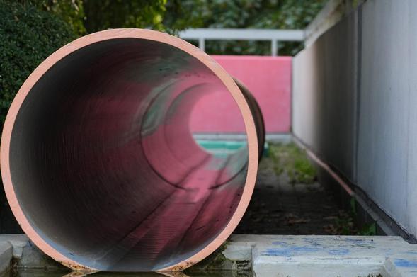 A very large pipe connects two small bodies of water on a concrete terrace. The rim of the pipe faces the camera and is a faded orange color. The inside of the pipe reflects the faded pink terrace parapet in the background. The ground is somewhat overgrown and there are more, bigger plants on the side and behind the parapet.