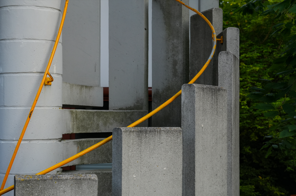 A freestanding spiral staircase made of concrete. Only the right half and the center pillar of the staircase are pictured, the left half is cut off. The concrete stairs and guard pieces as well as the orange hand rails all move upwards as the staircase goes on, giving a nicely structured picture that contrasts with the somewhat dirty and withered condition.