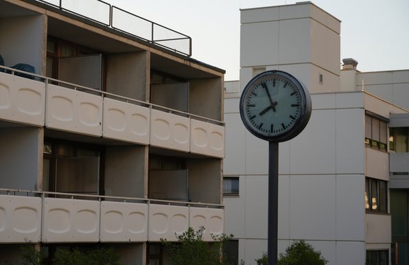 A station clock stands in front of an aged white slab concrete house covered in balconies. The slightly orange lighting suggests the sun might be about to set soon, and the clock confirms this, showing it's almost 8 pm.