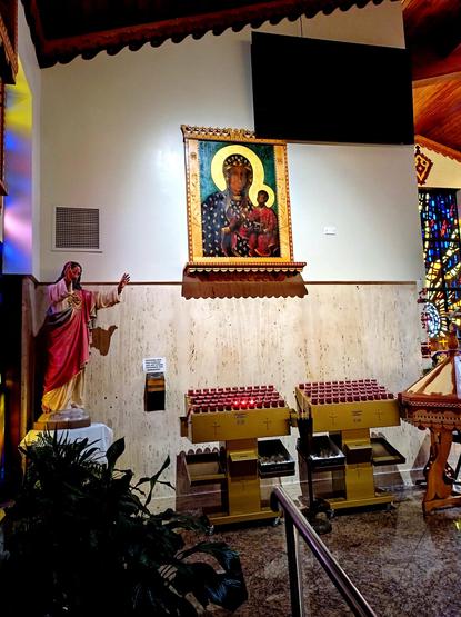 A photograph inside a church with an ancient style icon of the Virgin Mary and baby Jesus is above two tables of vigil candles. Three candles burn in the first row of a rack. A statue of an adult Jesus reaches out toward the icon. Multiple colors shine on a wall from a stained glass window on the left. Another stained glass window is visible on the right.