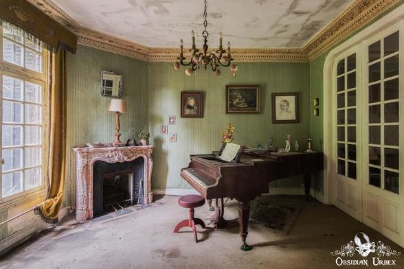 An abandoned room with a grand piano, a fireplace, framed pictures on green walls, a chandelier, and dusty furniture. Light enters through tall windows with gold curtains.