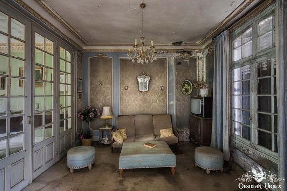 An abandoned, vintage living room with faded wallpaper, worn furniture, a small chandelier, and a dusty TV. Light enters through large windows and glass-paneled doors.