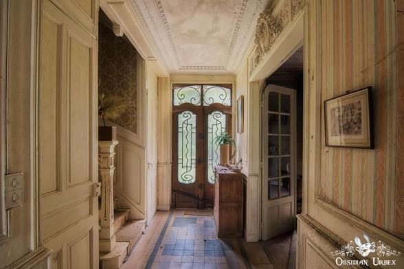 A hallway in an old, abandoned house with ornate wooden doors, faded wallpaper, tiled floor, and a small cabinet under a window.