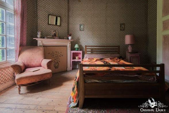 A vintage bedroom with floral wallpaper, an armchair, a wooden bed with a colorful blanket, a fireplace, and a window letting in natural light.