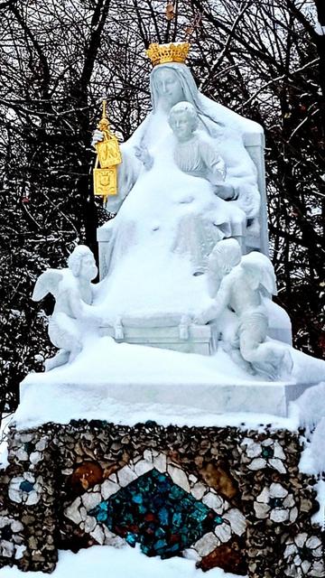 A statue of Mother Mary and baby Jesus sitting on a throne is covered with snow. The Mary figure has a gold crown and is holding a gold scapular. There are cherubs kneeling at the sides of the throne. The base is made of small stones and an inlaid aquamarine diamond shape made of stones. The diamond shape is outlined in white.
