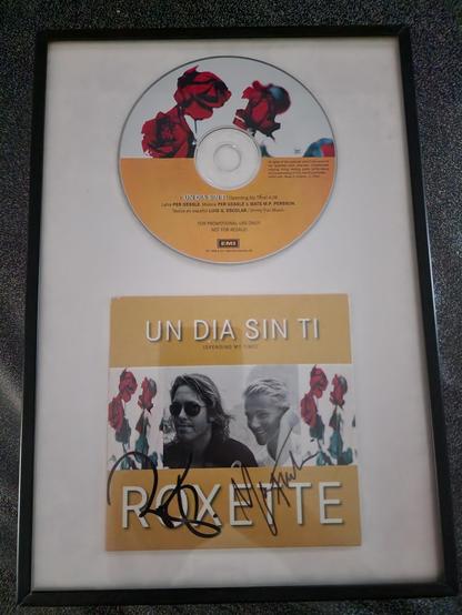 A neatly framed display showcases a rare Roxette demo single titled “Un Día Sin Ti (Spending My Time)”. In the upper half of the frame sits the promotional CD, its artwork featuring vivid red roses against a soft gradient of white and warm yellow. Beneath it rests the matching sleeve, also in yellow tones, with the title printed in bold white letters. The artwork shows Per Gessle wearing dark sunglasses beside Marie Fredriksson, both smiling gently, framed by tall red roses on either side. The sleeve is signed by both Per and Marie in bold black marker, making it a cherished collectible. The frame has a simple black border and white backing, highlighting the item’s clean, elegant presentation. This promo single was won by Cynni in a raffle held by the Roxette fanclub back in the day, making the piece not only rare but personally meaningful.