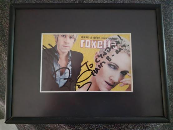 A black, matte-framed display holds a vibrant Roxette promotional postcard from the “Have a Nice Day” era. The postcard features a split portrait of Per Gessle on the left, wearing a dark jacket over a light shirt, gazing confidently at the camera, and Marie Fredriksson on the right, leaning forward with a striking, intense look against a sunny yellow background. At the top, the postcard reads “Have a Nice Postcard – Roxette” in playful, bold lettering. Both Per and Marie have signed the card in black marker, with Marie’s message reading “To Cynni – Happy B-Day!” in her distinctive handwriting. The autographs curve across their portraits, giving the postcard a personal and cherished touch. The frame’s dark border contrasts with the bright colors of the card, making the signatures stand out. This treasured keepsake was a birthday gift from Cynni’s best friend, who met Per and Marie during their promotional visit to the Netherlands and had the card signed especially for her.