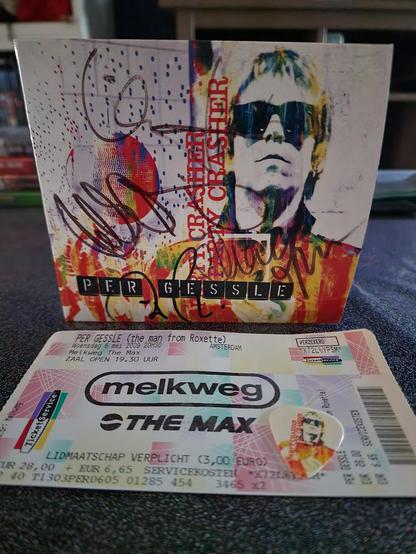 A vibrant, autographed Per Gessle CD sleeve stands upright on a dark surface. The cover, from his Party Crasher era, features a bold, colorful collage of Per wearing dark sunglasses, surrounded by splashes of red, yellow, blue, and abstract dotted patterns. Several signatures sweep dramatically across the artwork in black marker — Per’s autograph along with those of a few band members, all added after the concert at the Melkweg in Amsterdam on May 6, 2009. In front of the sleeve lies the original concert ticket, printed in soft pastel hues with the “Melkweg – The Max” logo prominently displayed. The ticket confirms the show as “Per Gessle (the man from Roxette)” and lists the date, venue, and opening time. Resting beside the ticket is a small white guitar plectrum featuring Per’s stylized portrait from the same album artwork. The arrangement captures a treasured memory of the night Cynni saw Per live and brought home both signatures and a plectrum from the gig.