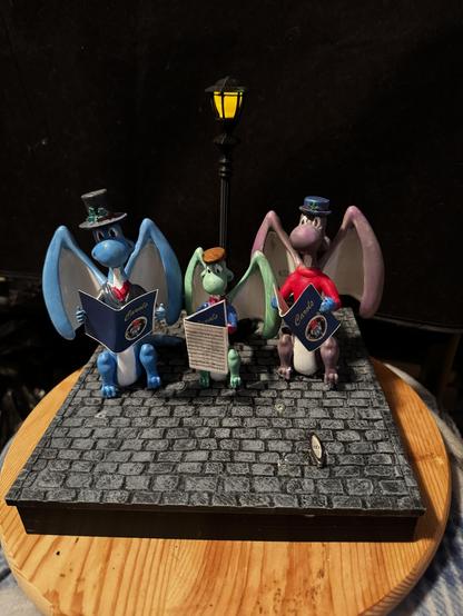 A 3D Printed holiday scene of 3 tiny dragons - 2 adults and a child, holding caroling books and standing in front of an old fashioned lamp post - they are wearing Victorian style caroling outfits
