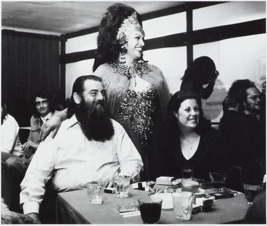 A black and white photograph captures a lively social scene where individuals are gathered around tables. The focus is on two main subjects: in the foreground, an individual with long hair seated at a table adorned with glasses of beverages; in the background stands another person wearing what appears to be elaborate drag attire featuring glittery decorations, suggesting this may have been taken during or after a performance event like a cabaret show. Surrounding individuals are engaged and attentive, some leaning forward while others smile broadly, indicating enjoyment. The setting suggests an indoor venue with natural light filtering through windows in the background, typical of establishments that host social events.