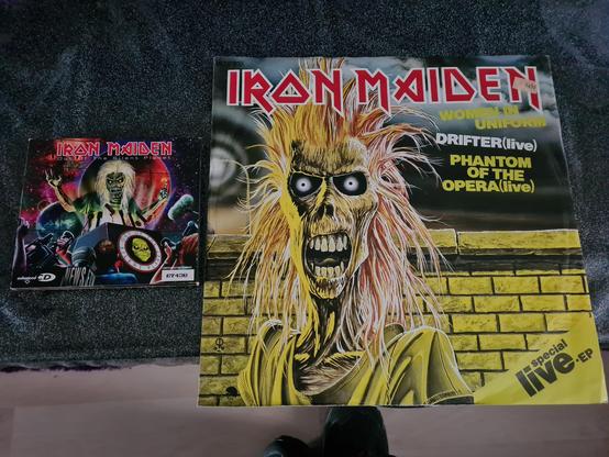 The image shows two Iron Maiden releases displayed side by side on a dark, textured surface. On the right lies a large 12-inch vinyl single featuring the band’s iconic mascot, Eddie, in a wild, early-era design: his hair is spiked and chaotic, his eyes glowing white, and his teeth bared in a menacing grin. Behind him is a yellow brick wall, with bold text listing the tracks “Women in Uniform,” “Drifter (live),” and “Phantom of the Opera (live).” The vivid artwork is dominated by reds, yellows, and blacks, capturing Iron Maiden’s classic heavy-metal aesthetic. On the left is a smaller CD single for “Out of the Silent Planet,” showcasing a more modern sci-fi themed Eddie, illuminated in neon colors against a cosmic background filled with planets and swirling galaxies. Both covers highlight the evolution of the band’s artwork over the years, making the display a striking contrast between eras of Iron Maiden’s visual and musical identity.