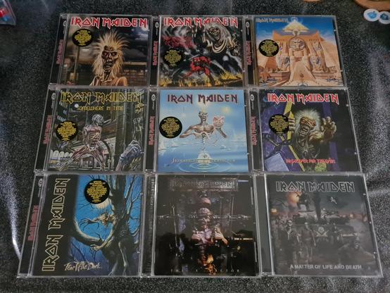 A neatly arranged 3×3 grid of Iron Maiden CD albums is displayed on a dark, textured surface, showcasing part of Cynni’s Iron Maiden collection. Each jewel case features the band’s iconic mascot, Eddie, in dramatically different styles across the eras. The top row includes Iron Maiden with its wild-haired street-punk Eddie; The Number of the Beast with Eddie towering over flames; and Powerslave, dominated by an Egyptian-themed Eddie carved into a massive temple façade. The middle row shows the futuristic cyborg Eddie of Somewhere in Time; the serene, icy blue landscapes of Seventh Son of a Seventh Son; and the snarling graveyard Eddie from No Prayer for the Dying. The bottom row features the shadowy, tree-dwelling monster Eddie of Fear of the Dark; the dark, industrial realism of The X Factor; and the war-torn battlefield imagery of A Matter of Life and Death, with Eddie leading a ghostly platoon. Together, the albums form a vivid timeline of the band’s evolving artwork and sound.