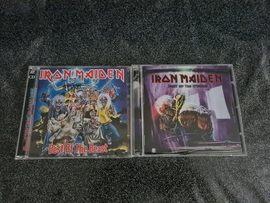 Two Iron Maiden compilation CDs lie side by side on a dark, speckled surface, showcasing highlights of Cynni’s collection. On the left is Best of the Beast, a 2-CD set with a chaotic and colorful collage of multiple versions of Eddie, the band’s mascot, from different eras. They surge forward as a terrifying crowd—ranging from skeletal to cyborg—to create a dramatic tribute to Maiden’s classic years. The band’s bold red logo stretches across the top, with the title Best of the Beast in striking red lettering below. On the right is Best of the B’Sides, featuring a darker, moodier cover: Eddie crouches inside a dim, bluish-purple space, spray-painting “Maiden Rule” onto a wall while glancing back over his shoulder with a wicked grin. The title sits above him in sharp red type. Together, the two compilations celebrate both Iron Maiden’s greatest hits and their lesser-known gems, capturing the band’s enduring creativity and iconic artwork.
