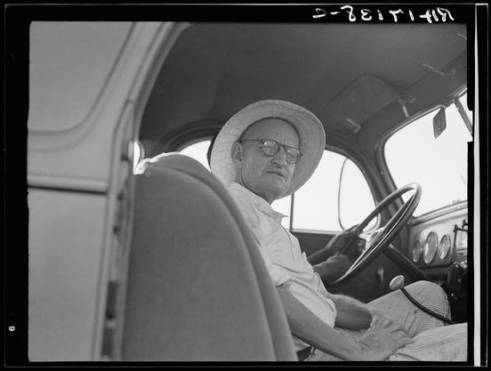 The image is a black and white photograph of an older man sitting in the driver's seat of a vintage vehicle. He wears round glasses, has gray hair, and dons a wide-brimmed hat that extends down to his neck. His expression appears focused as he gazes off into the distance while grasping the steering wheel with one hand. The interior of the vehicle is visible behind him; it features a simple dashboard with gauges and dials indicative of early automotive design, along with various other controls on the console.

The photograph captures an intimate moment in what seems to be a personal or historical context related to farming. A handwritten caption at the top right corner reads "D-ZEIT I+I," which could potentially relate to German words meaning time and date (Zeit für Zeit), but its significance is unclear without additional information.

In terms of background, there's no explicit depiction of an external environment; instead, we see only a glimpse through what appears to be the side window or door of another vehicle. The monochromatic nature of the photo adds a timeless quality and suggests it could have been taken in mid-20th century America during a period when rural life was deeply connected with agricultural work.

The accompanying information provided mentions "A tractor pioneer of the Mississippi Delta," indicating that this man may be associated with pioneering advancements or practices related to farming. The hist [...]