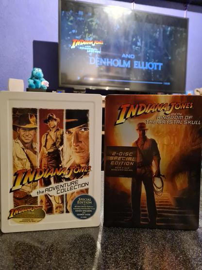 Two steel-case editions of the Indiana Jones films stand side by side on a dark, reflective surface. On the left is Indiana Jones: The Adventure Collection, featuring a collage of Harrison Ford as Indy in three iconic poses—hat tilted, whip in hand, dust-covered and determined. The artwork is framed in a white metal case with warm sepia tones, and a blue “Special Edition” sticker adorns the lower right corner. On the right is the steel-case edition of Indiana Jones and the Kingdom of the Crystal Skull, presented in deep oranges and browns. The cover shows Indy standing heroically at the entrance of a glowing temple staircase, his silhouette sharp against the fiery light behind him, holding his signature whip. A “2-Disc Special Edition” label sits near the center. In the background, a TV screen plays the first Indiana Jones movie via Netflix, showing the opening credits with Denholm Elliott’s name appearing over a jungle scene. The purple-lit room adds a cozy movie-night atmosphere.