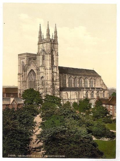 The image shows a historic church building with Gothic architectural features, including pointed arches and tall spires. The photograph appears to be an old postcard or print, given its sepia tone and the way it captures details of the structure and surroundings. In front of the church is a well-maintained garden area with pathways leading towards different parts of the grounds. Several trees are visible in the foreground, providing a green contrast against the stone facade of the building. The photograph also includes text that identifies the location as Bridlington Quay Priory Church and provides a reference number for cataloging purposes (10369).