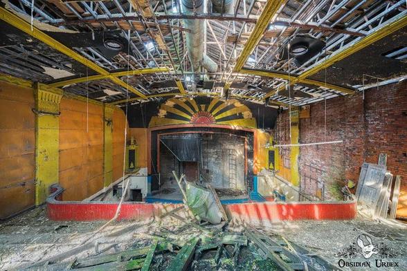 Abandoned theater with a damaged stage, exposed ceiling beams, debris on the floor, and faded yellow and red walls.