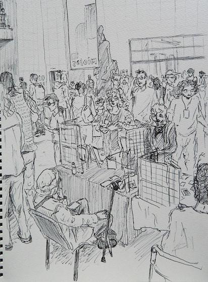 Black and white drawing made with a pen. It's from the perspective of the back of Ya's table, they are sitting on a chair, with their back to us, there's a cane to the side and a few grids on the table, where they hang their works. There's a lot of people walking around, a table to the right and artists standing on the left. We can also see the back of the statue of the previous drawing on the background and the bottom part of the banner.