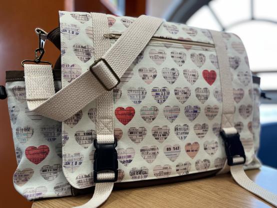 A messenger bag with a cream background and brown body canvas. The cream fabric is printed with repeating hearts of library due-date slips. Occasional hearts are red. It is extremely cute and makes one think “ca-chonka.”