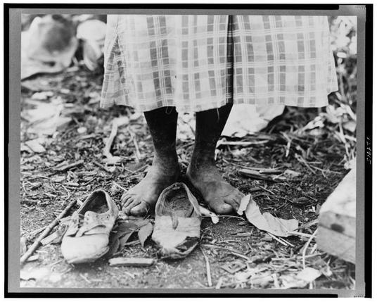 The black and white photograph shows a close-up of a person's feet standing on the ground with no shoes. The individual is wearing a plaid skirt or short, which reveals their legs down to just above the knees. Their bare skin appears wrinkled and weathered, suggesting they may be an elderly person or someone who has endured difficult conditions.

The person stands next to two discarded footwear: one shoe that seems worn out with some dirt on it, and a sockless foot wearing what looks like a torn piece of cloth. The ground is littered with debris such as leaves, twigs, and possibly small rocks or stones. There's also an object resembling the bottom part of another shoe next to the feet.

In the background, there are blurry shapes that may indicate more people or objects on the ground in what seems like a rural setting. This image evokes themes of poverty, neglect, or hardship faced by individuals who lack basic necessities such as shoes and clean living conditions. The photograph is credited with being taken near Clarksdale, Mississippi.

For context about Dorothea Lange's work during that period, she was known for documenting the effects of the Great Depression in America through her powerful photographs capturing human suffering and resilience.