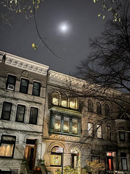 over an ornate urban brownstone rowhouse with festively lit windows, the moon shines through mist