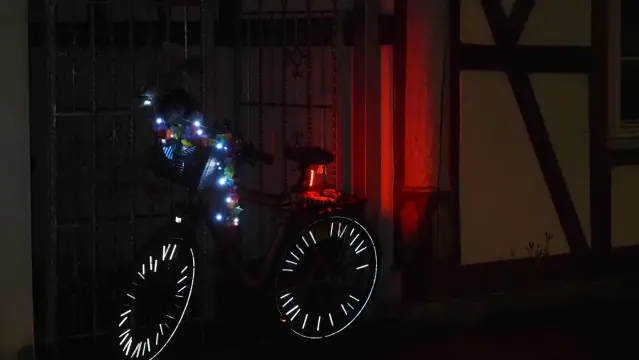 Fahrrad mit Lenkerkorb lehnt an einem Gitter. Die Lichterkette am Lenkerkorb leuchtet und die Speichenstick (es fehlen ein paar)  reflektieren