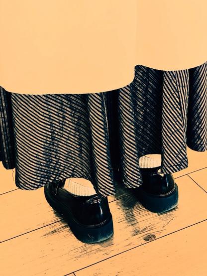From bottom to top: a wooden floor, black shoes, grey socks, black and white diagonally striped long dress, camel-coloured coat. From the back.