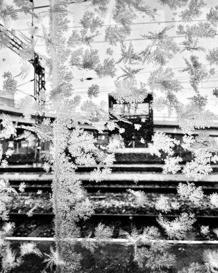 Frosty patterns cover a window, revealing blurred train tracks and infrastructure in the background, evoking a chilly, quiet atmosphere.