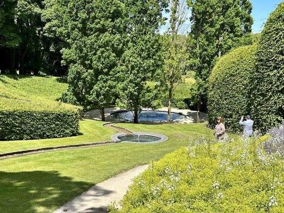 A landscaped garden features a small circular fountain at the centre of a narrow, gently curving water channel leading toward a larger round pond surrounded by trees. The neatly trimmed lawns are bordered by dense green hedges and flowering plants, with patches of yellow and white blooms in the foreground. Two people stand near the flowers, one holding a camera and the other observing the surroundings. Tall trees and thick hedgerows provide a natural enclosure, with sunlight casting soft shadows across the grass. The scene is calm, structured, and well-maintained, highlighting the contrast between open spaces and dense greenery.
