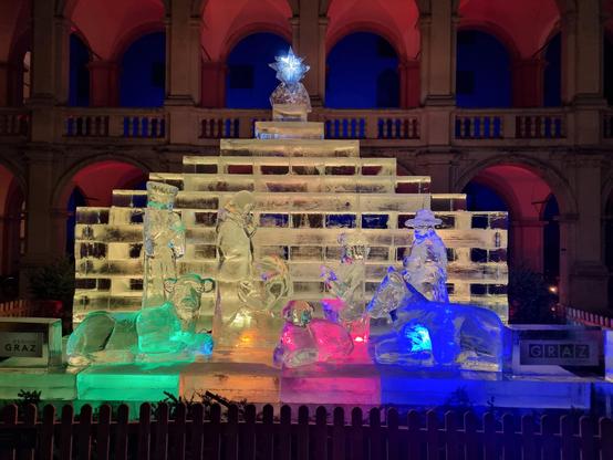 Eisfiguren der Krippe. Dahinter aus Eis eine Hütte vor beleuchteten Arkaden