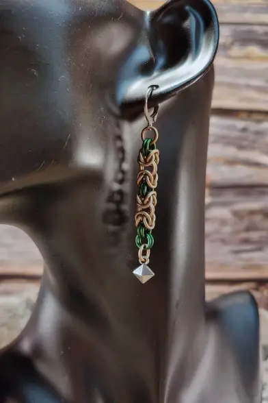 Boucles d'oreilles en maille, dans des couleurs taupe et vert forêt qui alternent. Au bout, un petit pendant de couleur argentée y est attaché, tel un rayon de lumière dans une clairière.