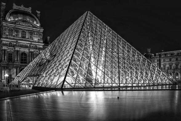 Schwarz Weiss Aufnahme der Glaspyramide im Zentralen Innenhof des Louvre Palastes in Paris. Links und rechts erkennt man die rückwärtige Fassade des Louvre Palastes. Die Pyramide ist von innen beleuchtet.