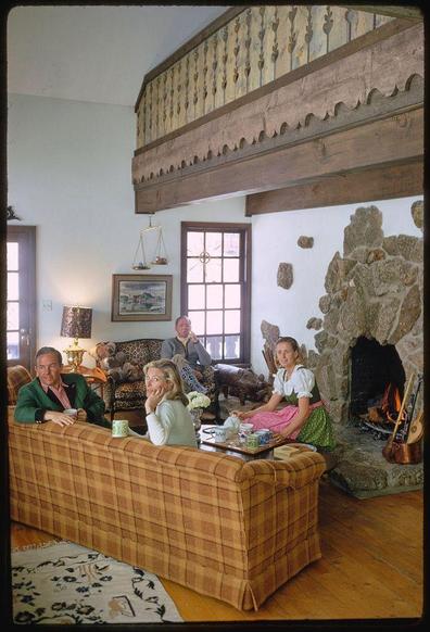 Image displaying a cozy interior scene, featuring four individuals situated across seating arrangements that include sofas and armchairs. The predominant color scheme comprises warm tones with touches of greenery from plants or textiles.

On the left side, an individual in a striking lime-green jacket paired with dark trousers occupies a sofa, facing towards two other seated people on a brown checkered couch; one is clad in traditional attire resembling Alpine-style dress while another wears casual clothing. The person wearing the distinctive outfit appears to be interacting playfully or posing for the camera.

In contrast, an individual sits alone at a separate table placed near a stone fireplace with its door open and flames visible inside. This setting also includes various items such as a guitar resting against the wall, a lampshade, artwork on display, and numerous windows allowing natural light to penetrate into the space.