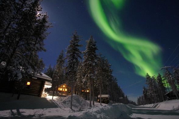 Das Bild zeigt eine Nachtszene in einer verschneiten Landschaft. Im Vordergrund befinden sich mehrere Holzgebäude, einige davon beleuchtet. Die Gebäude stehen zwischen hohen Nadelbäumen, die ebenfalls mit Schnee bedeckt sind. Am Nachthimmel befindet sich ein grünes Nordlicht, das in Bögen über den Himmel verläuft. Der Schnee bedeckt den Boden und die Dächer der Gebäude, und es gibt eine Lichtspur, die sich durch die Landschaft schlängelt.