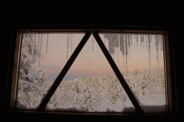 The image depicts a winter scene viewed through a window with a triangular frame. The window is partially covered with icicles hanging from the top, creating a triangular pattern. The view outside shows a snow-covered landscape with trees heavily laden with snow, indicating a recent snowfall. The sky is painted with soft pastel colors, suggesting either sunrise or sunset. The snow on the ground and trees is pristine and untouched, emphasizing the stillness of the scene. The overall color palette is dominated by whites and soft pastels, with the icicles adding a touch of gray.