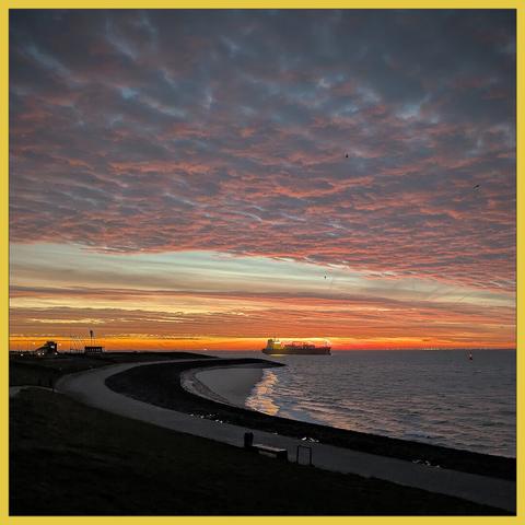 The clouds in the morningsky colors orange just before sunrise. A large ship stands in the center of the picture. In front small breaking waves, a curved dyke with a footpath.