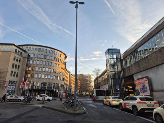View of the nearest crossroads, with sunlight reflected by the district town hall & library building, blue sky, the local tram station, a bus & some taxis waiting for clients.