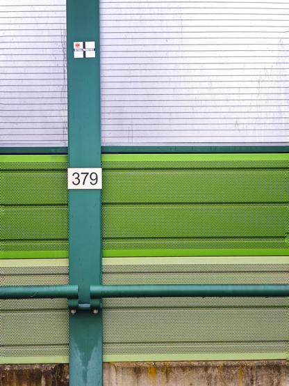Ausschnitt aus einer grau-grünen Schallschutzwand am Bahnhof. Auf einem Schild steht 379.