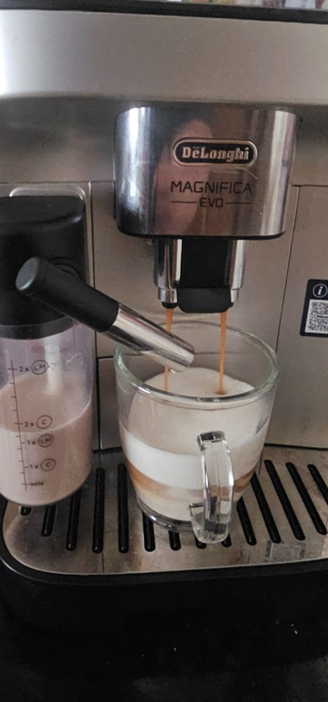 Kaffeeautomat macht Kaffee, eine durchsichtige Glastasse mit Hafermilchschaum in dem Kaffee reinläuft. Links der Hafermilchschaumbehälter ist haöbvoll.