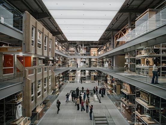 "Multi-story museum archive with visible storage collections on three levels. Glass-walled display cases and artifacts line both sides of wide corridors. Visitors walk through the central space examining exhibits, while natural light filters through a translucent ceiling. The industrial-style architecture features exposed beams and modern materials creating an open, transparent repository."
This appears to be a "visible storage" style museum facility, where collections are stored in an accessible, educational way rather than hidden in traditional closed archives.