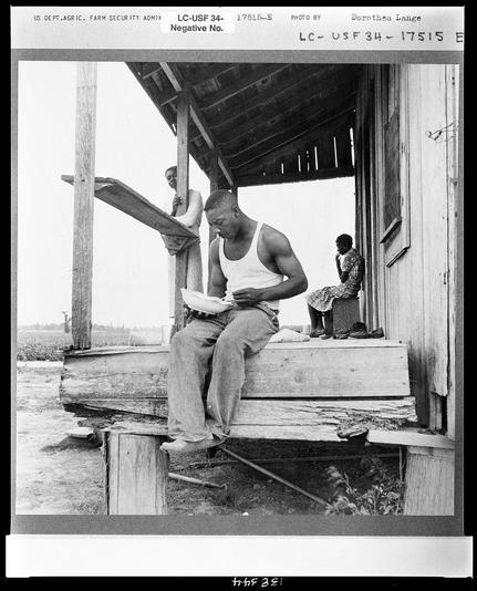 The black and white photograph features three individuals in an outdoor setting with a wooden structure, possibly a barn or shed. On the left side of the image sits a man wearing a sleeveless shirt reading from a document while resting his elbows on a makeshift ledge that supports him. Directly behind this individual stands another person who appears to be working at a task out of frame, and there's an open door leading into what might be a storage area or workspace. On the right side, a seated woman is visible in profile; she wears a patterned garment, possibly apron-style clothing over which hangs something like fabric or rags. Her posture suggests contemplation as her gaze seems directed off-camera to the left outside of view. The ground appears muddy and there are what could be plants growing near it on one side and another structure is barely visible in the far background with a hint of sky above, implying an open-air environment possibly related to agricultural work or rural setting. Textual information at the top includes identifiers such as "US DEPT.AGRIC. FARM SECURITY ADMIN," which indicates government association; photographer's name Dorothea Lange suggests historical context likely during her tenure documenting social issues like sharecropping in America; a reference number and date code, possibly for cataloging or research purposes within an archive collection, also suggest archival significance of the image.