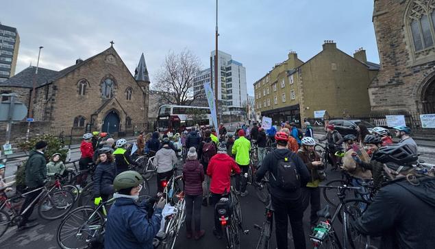 A large group of cyclists in variously coloured helmets, jackets and winter wear gather on an asphalt gyratory island flanked by roads, under a pale grey winter sky; they are being addressed by a blonde lady in a woollen hat using a microphone, while a double-decker bus goes past. Banners reading ‘The Causey’ - the name of the location and placemaking project - and ‘Critical Mass’, the name of the cycle ride - can be seen among the crowd. In the background, tall churches and university buildings loom.