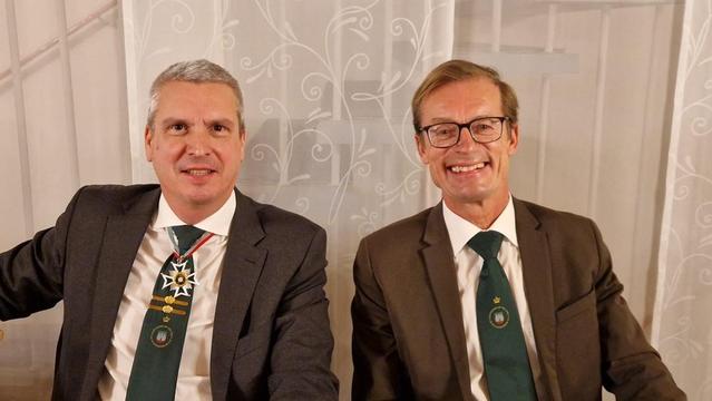 Zwei Männer in Anzug und Krawatte, mit Abzeichen, vor heller Wand.