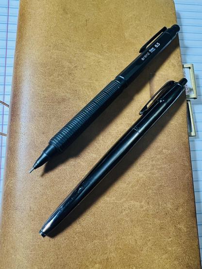 A Pentel Orenz Nero sitting next to a Fisher AG7-BTN. Black pen and mechanical pencil sitting on a leather notebook.