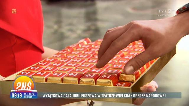 Zbliżenie na bombonierkę czekoladek z logo Orlen trzymaną przez hostessę ubraną w czerwoną - a jakże! - sukienkę, podczas gdy czyjaś ręka sięga po jedną z tych czekoladek. Źródło: TVP2, Pytanie na Śniadanie, 30.11.2025 r., godz. 09:19.