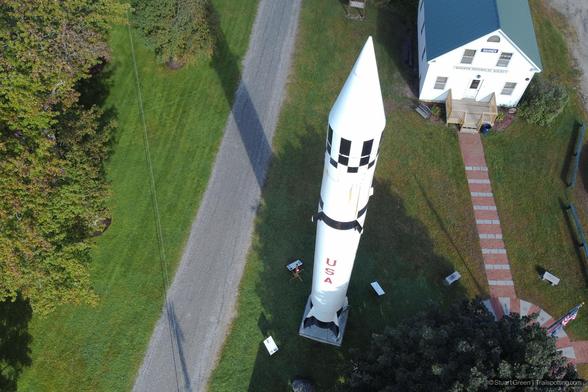 Aerial view of a historical site featuring an authentic Redstone rocket, painted white with black checkered bands and red 'USA' lettering, mounted on a concrete base amid green grass and trees. Informational plaques surround the rocket. Nearby stands a white building with a green roof labeled 'WARREN HISTORICAL SOCIETY' connected by a brick pathway. A gravel road runs alongside the scene.