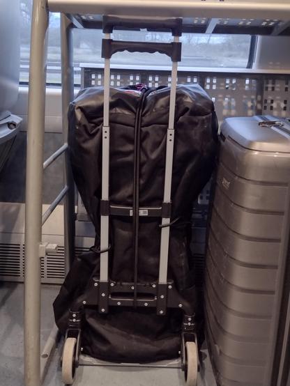 Eine robuste schwarze Reisetasche, etwa 1,10m hoch, ist festgezurrt auf einen Alu-Sackkarren-Gestell.