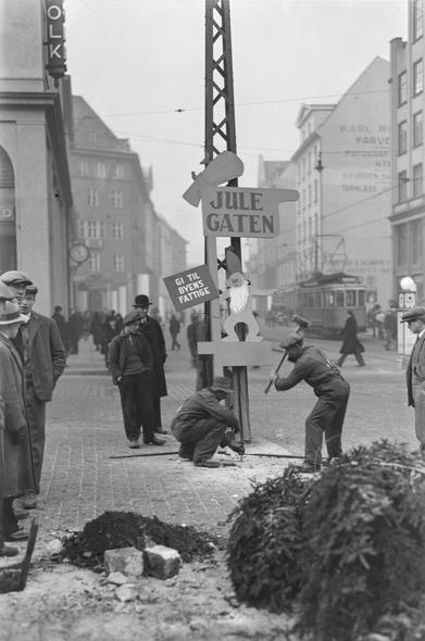 Black and white image. Two men, one with a sledge hammer, working signs and bolts into ground. People looking at them. Streetscape in background.