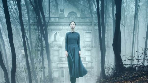 Gothic Horror film The Lodgers: Rachel (Charlotte Vega) stands in a long plain dark dress in front of a mist-shrouded house, walking through the woods in front of it. The trees in the foreground are a stronger colour than the washed-out, ghostly house in the background, and Rachel herself is pale and her clothes stand out like the trees, as if she is fighting to be real, but is herself an ethereal figure, caught between the natural world and the phantom world. Behind her, one of the upper windows of the house is open, and the white curtains are billowing outside. The rest of the house is shuttered and locked, with white blinds and curtains covering every other window.
